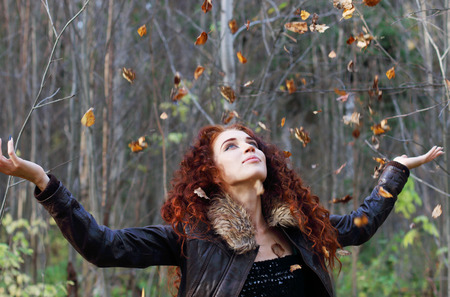 Beautiful woman with curly hair in leather jacket throws up dry leaves in sunny autumn forestの写真素材