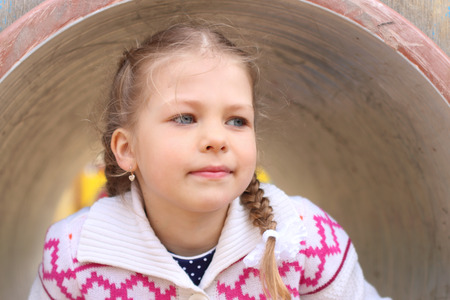 Pretty little girl peeks up of pipe on playground at summer day and looks awayの写真素材