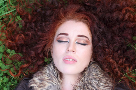 Pretty girl with red curly and closed eyes hair lies on green clover field, close upの写真素材
