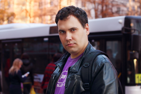 Man in leather jacket stands on street near bus stop in city, shallow dof