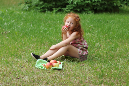 Little girl with red hair eats apple on lawn in green summer park, shallow dofの写真素材