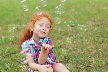 Little girl with red hair poses on grass among bubbles in summer park, shallow dofの写真素材