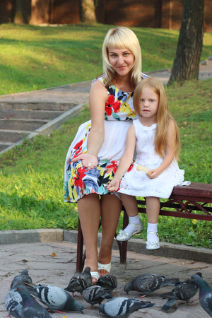 Pregnant mother and little daughter feed pigeons in summer sunny parkの写真素材