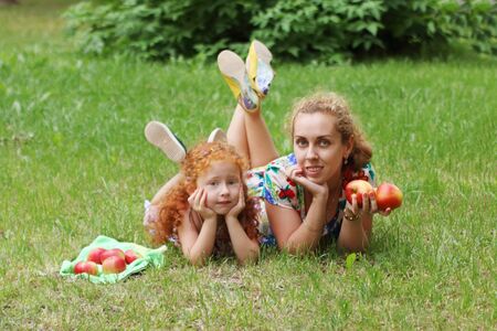 Cute little girl and her mother lie on lawn with apples in green summer park, shallow dofの写真素材
