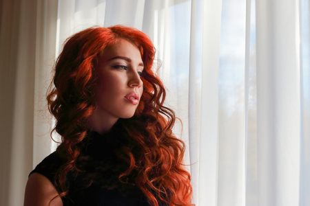 Young woman with beautiful curly hair standing at the window and gently looking forwardの写真素材