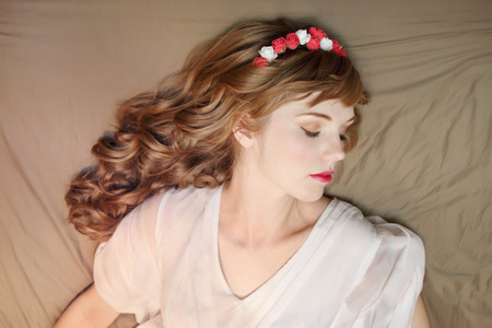 Dreaming girl in roses wreath and dress with curly hair lies on bed with closed eyesの写真素材
