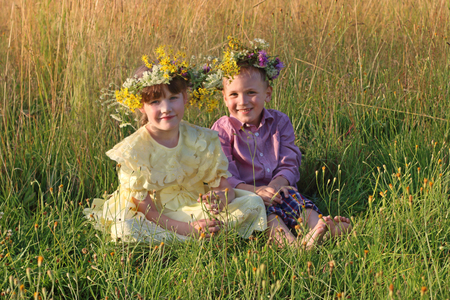 Happy little barefoot boy and girl in wreaths sit in dry grass at summer eveningの写真素材
