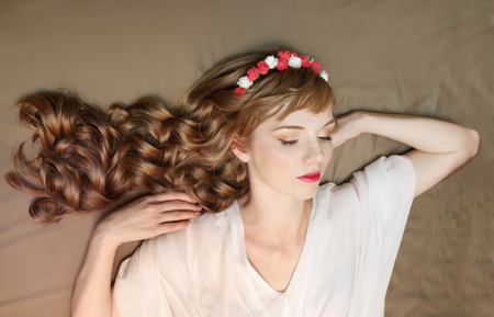 Cute dreaming girl in roses wreath and dress with curly hair lies on bedの写真素材