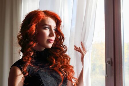 Cute red-haired girl in a beautiful lace black dress standing at the window and looks into the distanceの写真素材