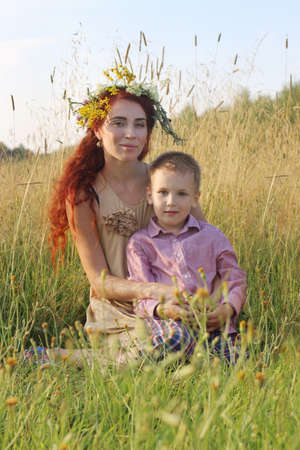 Happy woman in wreath and little son sit in dry grass at summer eveningの写真素材