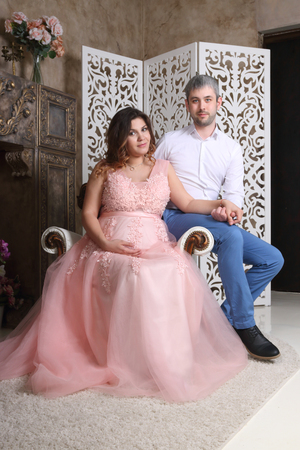 Man and pregnant woman in pink pose on baroque armchair in studioの写真素材