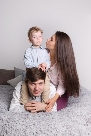 Happy father, mother, little son lies on grey bed in cozy bedroom, focus on man, woman kisses kidの写真素材