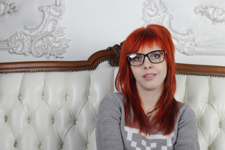Pretty girl with red hair in glasses sits on white sofa and smiles in white roomの写真素材