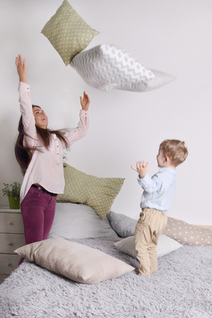 Young woman, little son throw pillows on grey bed in bedroomの写真素材