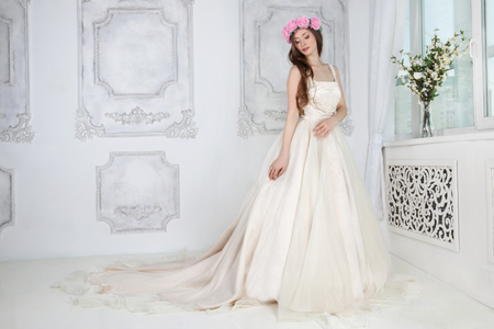Beautiful woman in pink roses wreath and in long dress poses in white studioの写真素材