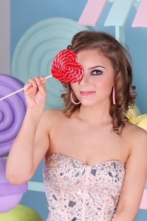 Cute curly woman hides her eye behind red lollipop as heart in studioの写真素材