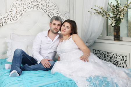 Young pretty pregnant woman in long dress and man poses on white bed in roomの写真素材