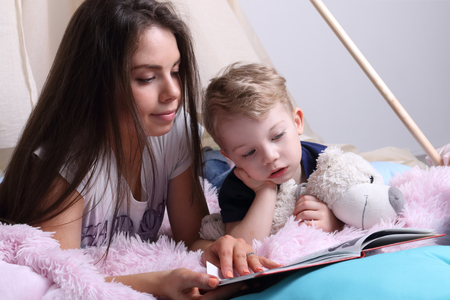Young mother reads book for her little cute son with toy among pillowsの写真素材
