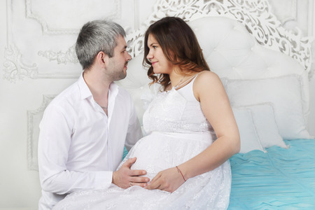Pregnant woman in dress and man look at each other on white bed in roomの写真素材