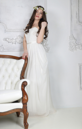 Young cute brunette in wreath and in white dress poses near couch in studioの写真素材