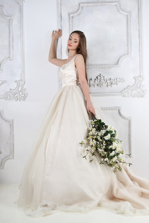 Young woman in long dress holds white flowers in white studio near wallの写真素材