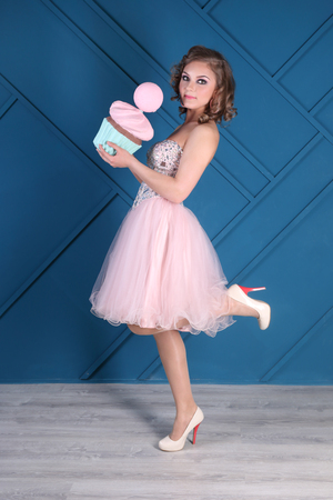 Pretty curly woman in pink dress and heels holds big cupcake in blue studioの写真素材