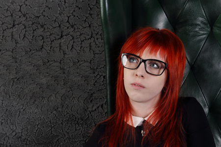 Woman in glasses and black dress sits in leather armchair and dreams in black studioの写真素材