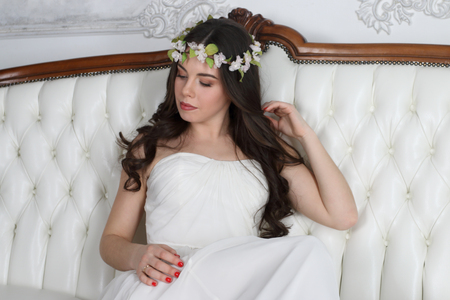Young cute brunette in wreath and in white dress poses on couch in studioの写真素材