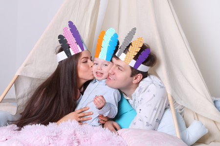Father, mother, little son pose in wigwam with indian feathers in studio, woman kisses boyの写真素材