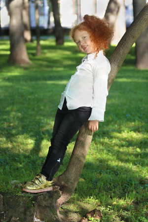 Little girl with red hair in white shirt poses near tree in summer green parkの写真素材