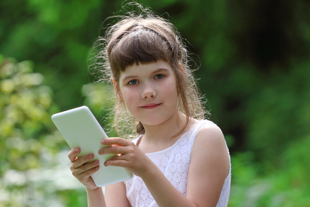 Little smiling girl in white dress holds white tablet pc in park at summer dayの写真素材