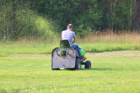 Lawn mower works on big green field near forest at summer day, back viewの写真素材