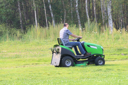 Lawn mower works on big green field near forest at summer dayの写真素材