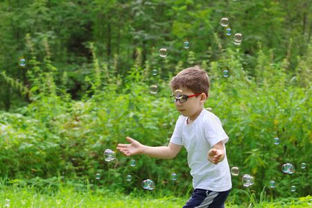 Happy little boy in glasses catches soap bubbles in summer green forest