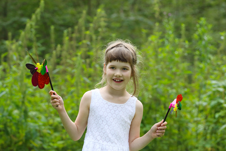 Little girl in white holds two windmills and smiles in summer green forestの写真素材