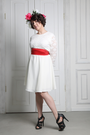 Beautiful young brunette woman in white dress with flower wreath in her hair posing near white door in photo studioの写真素材
