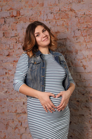 beautiful pregnant woman in striped dress, jacket is standing near brick wall in studio, palm-hearted gestureの写真素材