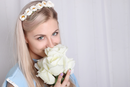 Beautiful young blonde woman with clean skin and flower wreath in her hair smelling bouquet white rose and smileの写真素材