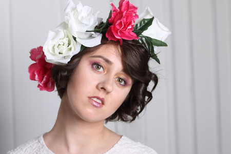young brunette woman with clean skin, make-up and flower wreath in her hair stands near white door, head on one sideの写真素材