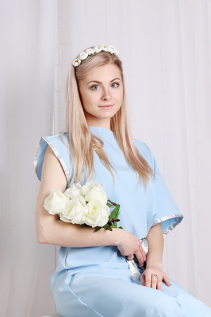 Beautiful young blonde woman with clean skin and flower wreath in her hair near curtain, roses greekの写真素材