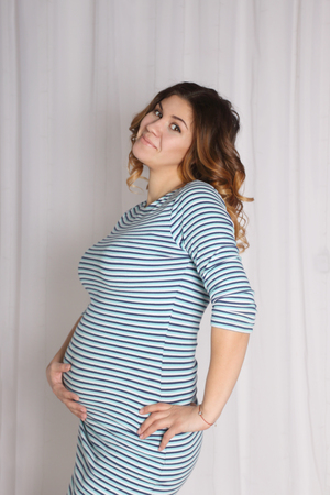 young pregnant woman in striped dress posing near white curtains in studio, hand lies on bellyの写真素材