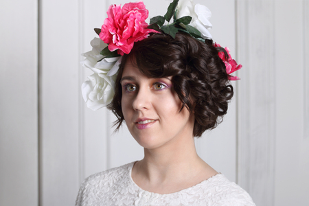 young brunette woman with clean skin, make-up and flower wreath in her hair stands near white door, look throwの写真素材