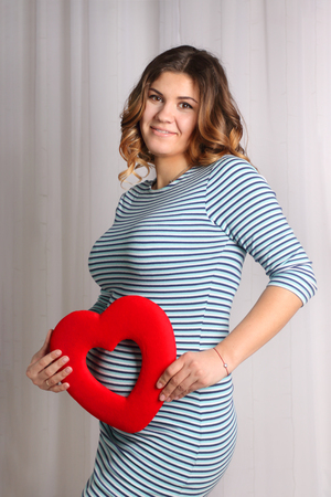 young pregnant woman in striped dress posing near white curtains in studio, holds red heartの写真素材