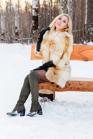 Beautiful blonde in fur coat, boots poses on wooden bench outdoor at winter day in parkの写真素材