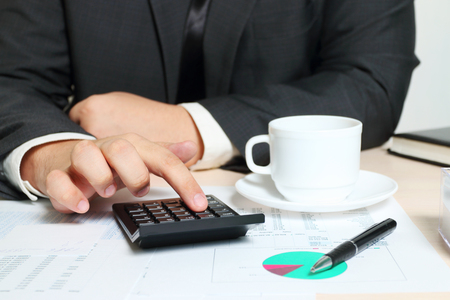 Hands of calculating businessman sitting at table with cup, documents, diagramsの写真素材