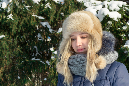 Sad young woman in fur hat looks down near green fir tree outdoor in sunny winter dayの写真素材