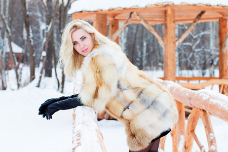 Beautiful blonde in fur coat, leather gloves poses outdoor at winter day in park with arborの写真素材