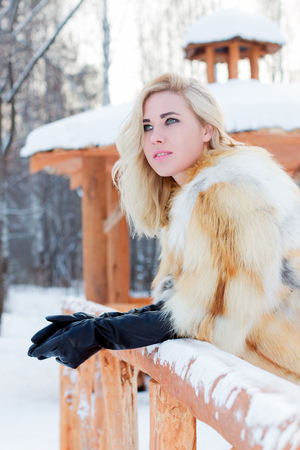 Pretty blonde in fur coat, leather gloves looks away outdoor at winter day in park with arborの写真素材