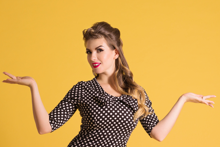 Pretty young woman in retro black dress with hairdo stands in yellow studio, pin up styleの写真素材