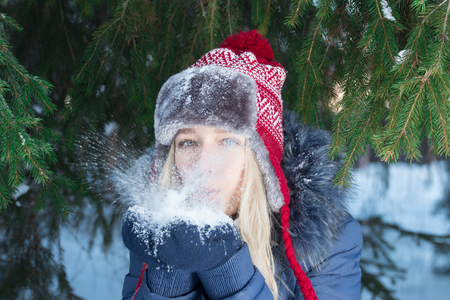 Girl in ear-flap hat blows out snow near green fir tree outdoor in winter dayの写真素材
